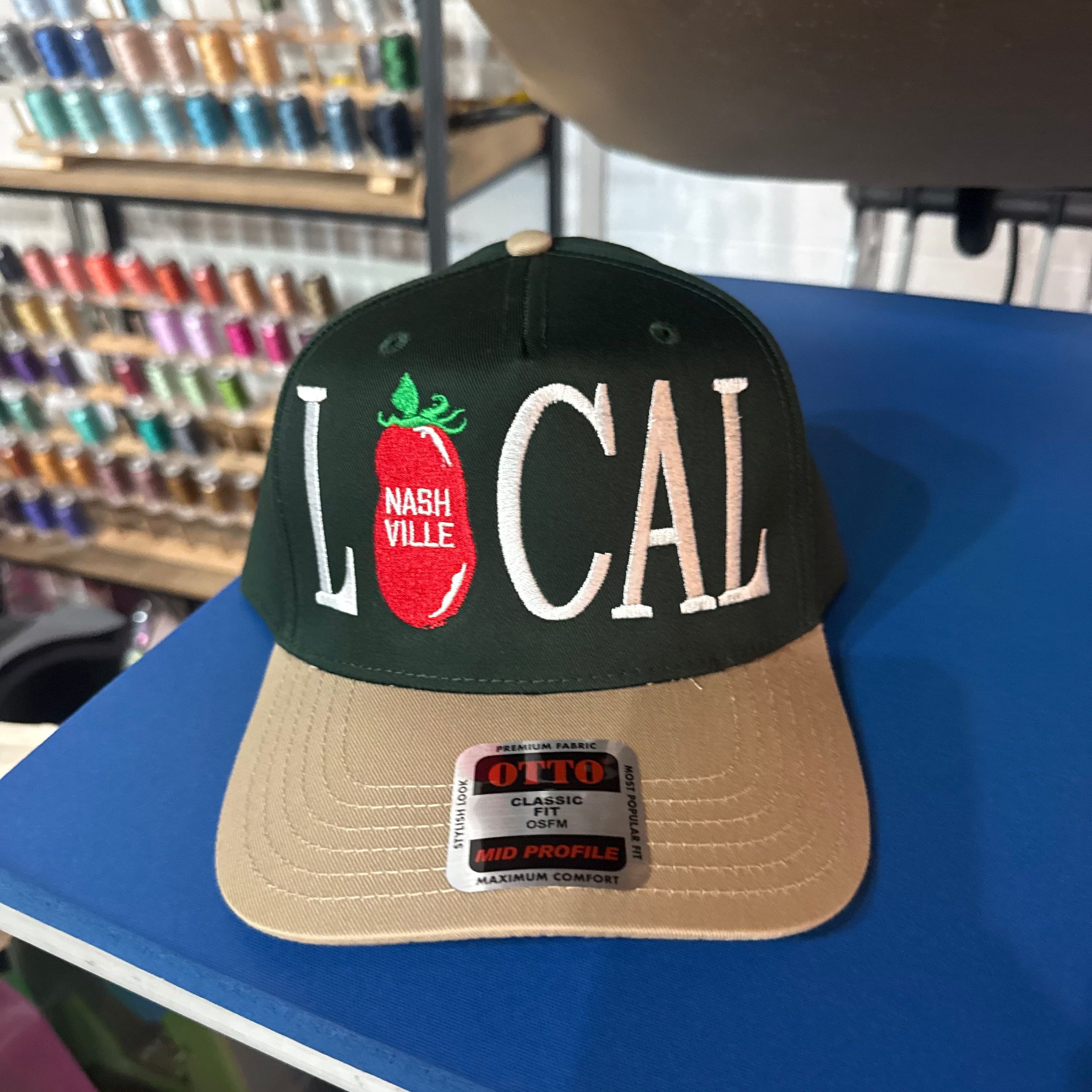 A dark green and tan snapback cap with 'LOCAL' and a tomato graphic on the front, displayed on a blue table with a Otto Cap brand sticker visible.