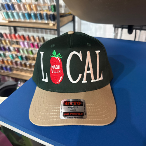 A dark green and tan snapback cap with 'LOCAL' and a tomato graphic on the front, displayed on a blue table with a Otto Cap brand sticker visible.