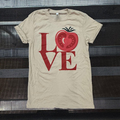 A beige short-sleeve t-shirt with a red graphic spelling 'LOVE' where the 'O' is a sliced tomato, laid flat on a black metal rack.