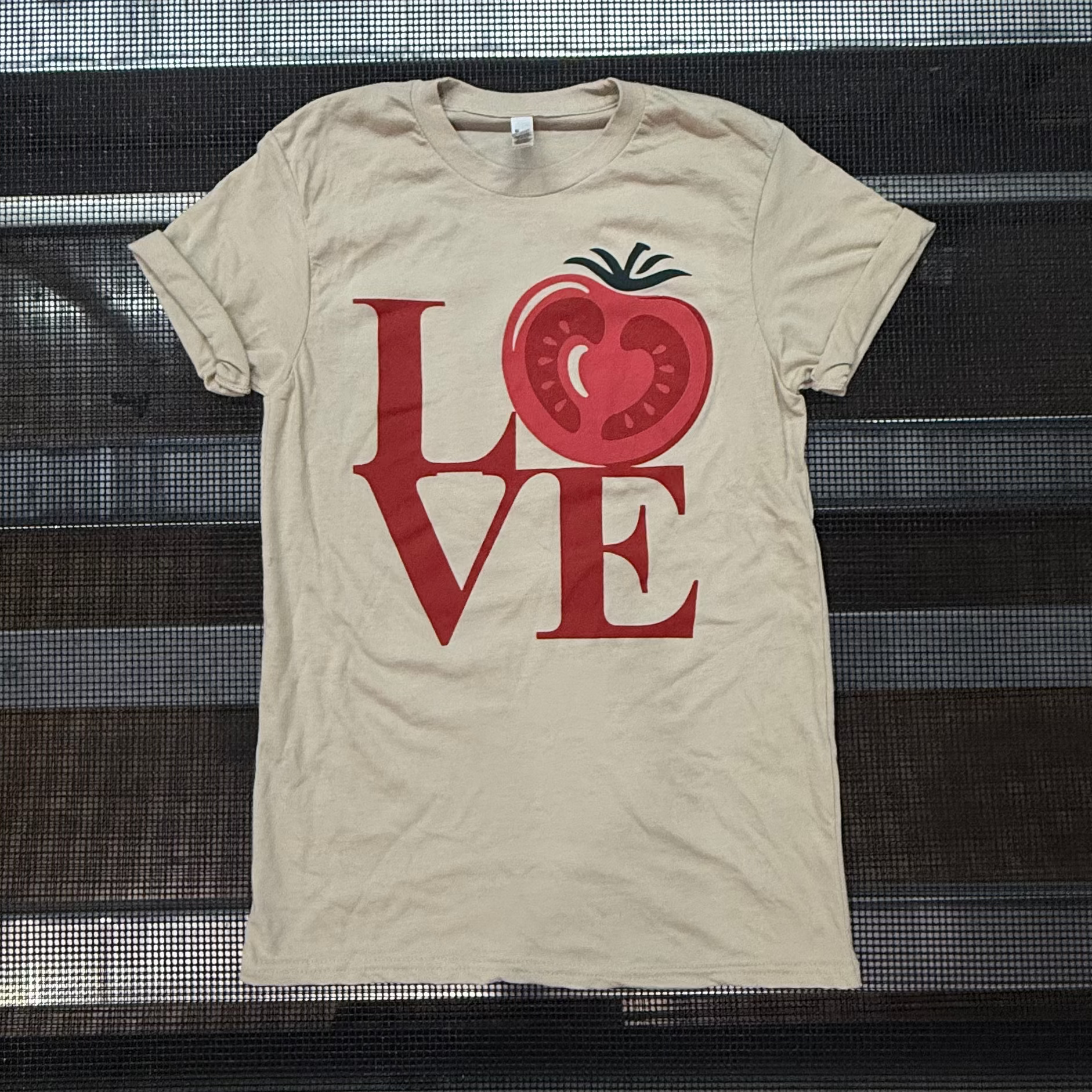 A beige short-sleeve t-shirt with a red graphic spelling 'LOVE' where the 'O' is a sliced tomato, laid flat on a black metal rack.