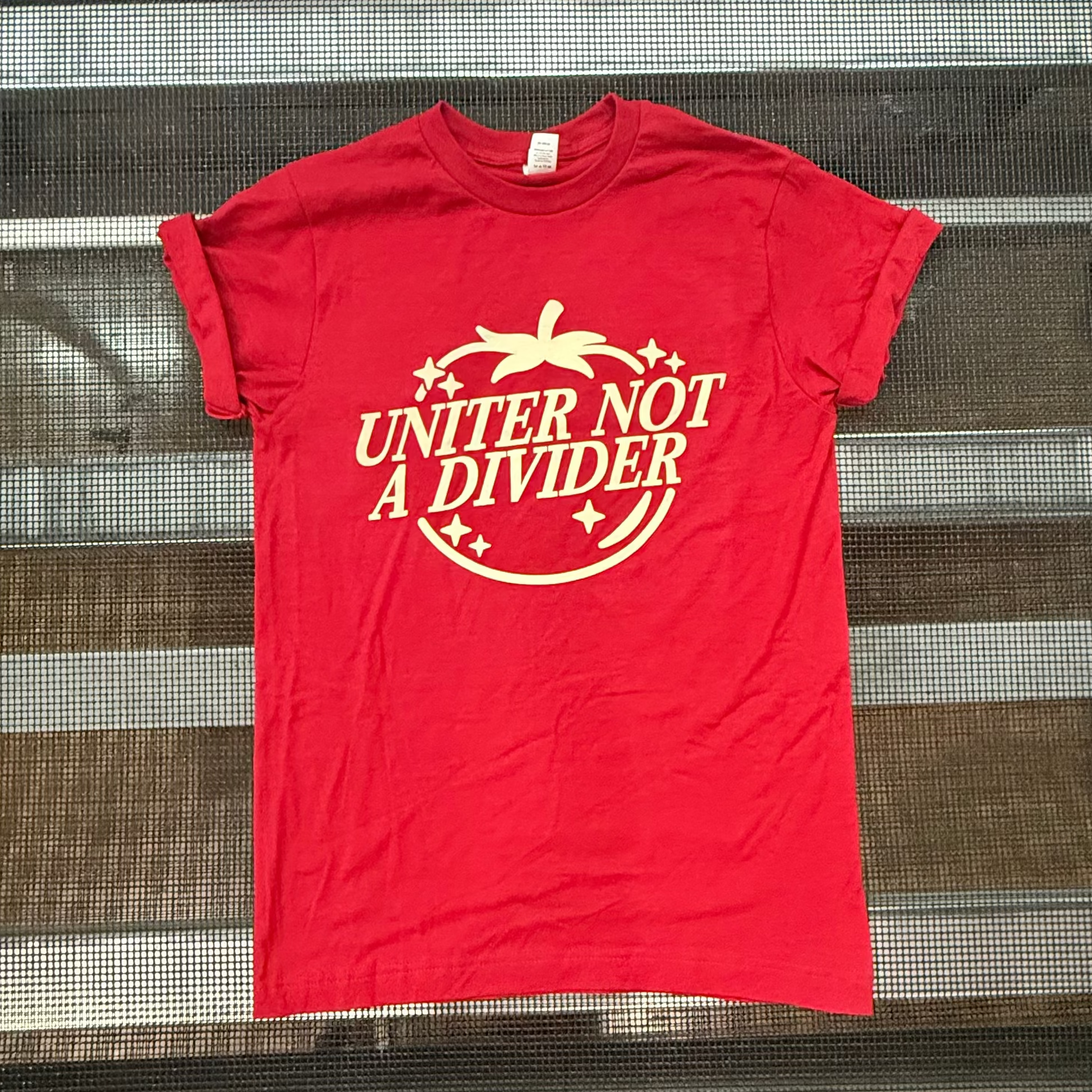 A red short-sleeve t-shirt with a white tomato graphic reading 'UNDER NOT A DIVIDER' , laid flat on a metal grid surface.