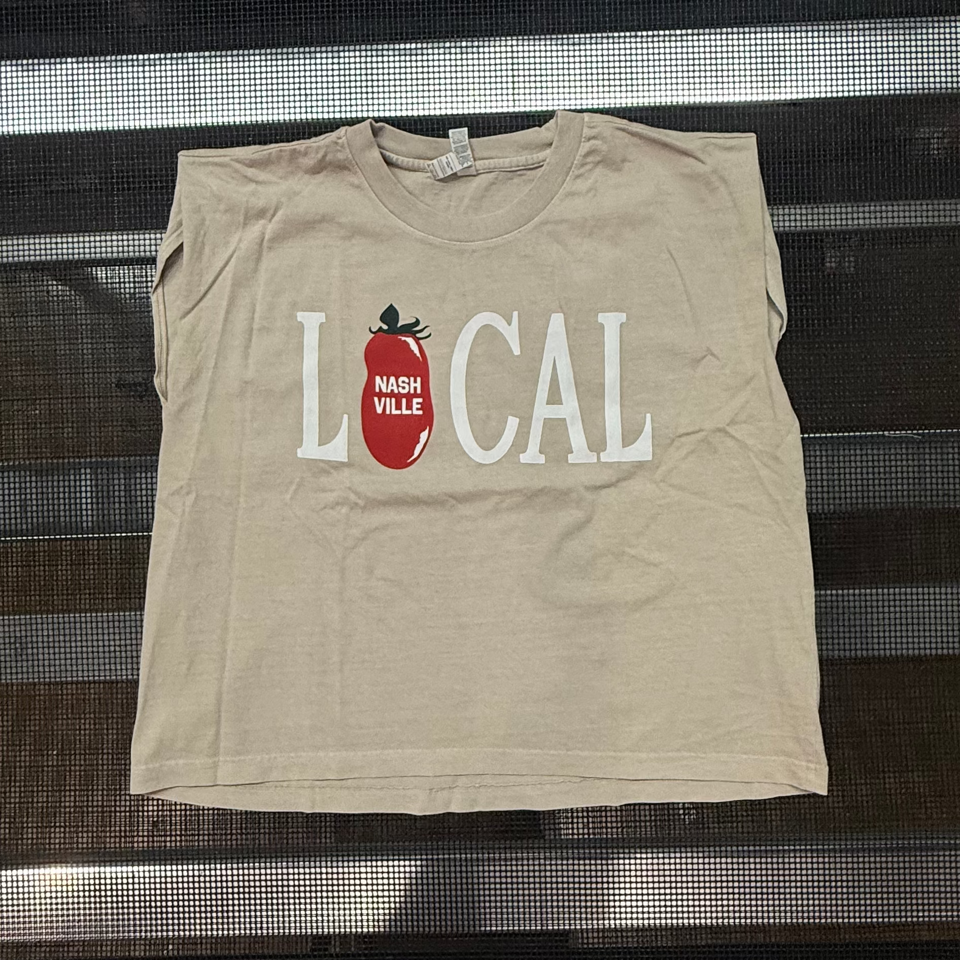 A beige sleeveless crop top with a graphic reading 'LOCAL' in white letters and a red tomato illustration, displayed on a dark metal rack.