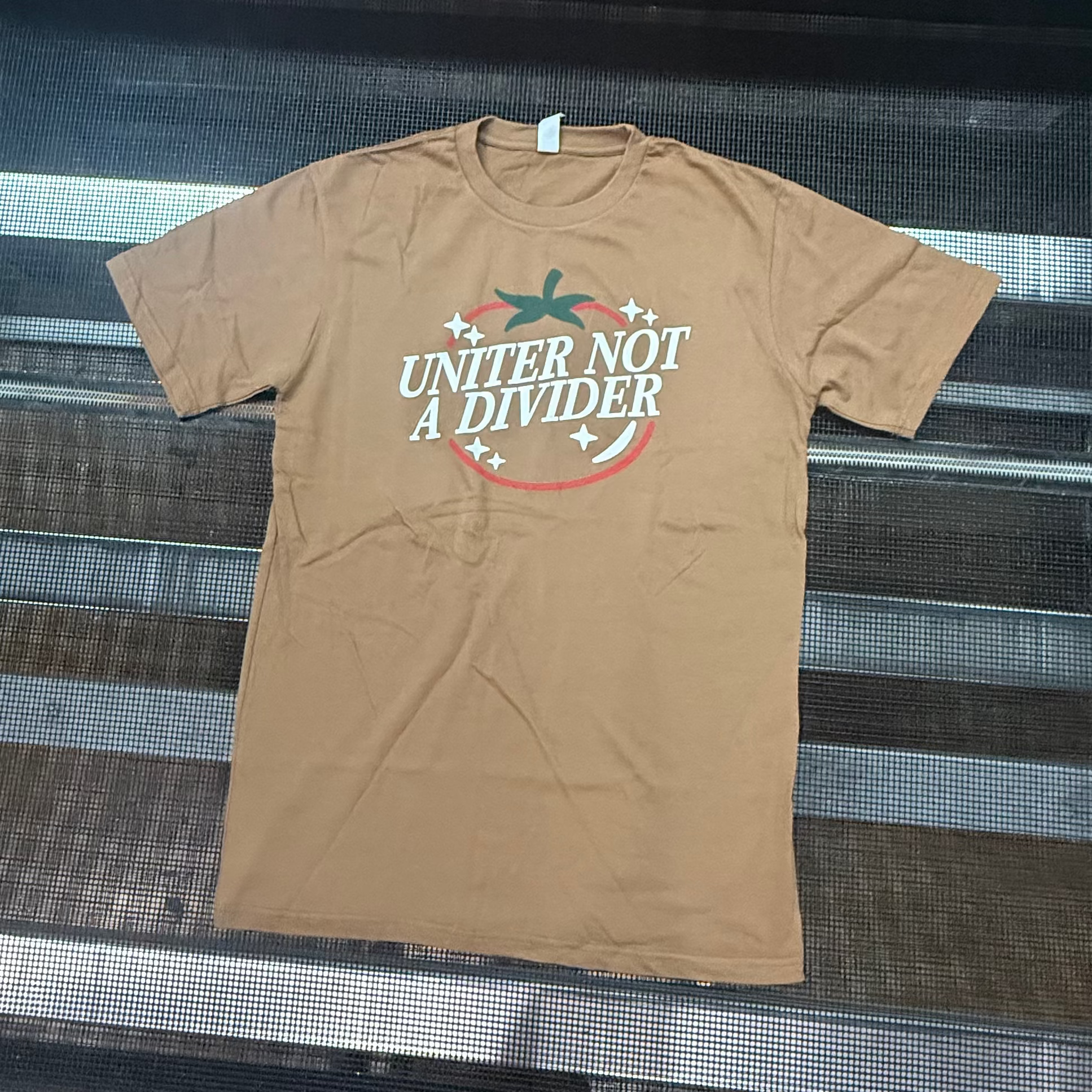 A brown crewneck t-shirt with a 'UNITE NOT A DIVIDER' graphic print, laid flat on the bed of a dryer.