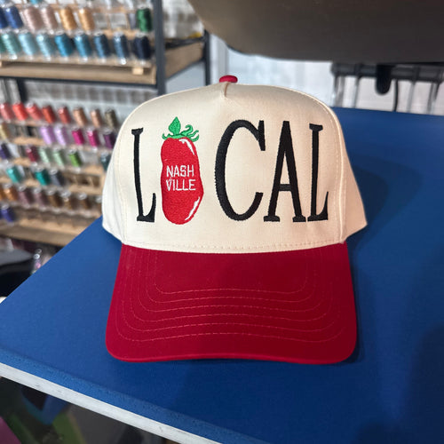 A red and cream trucker-style baseball cap with 'LOCAL' and a red tomato graphic featuring 'NASHVILLE' embroidered on the front, displayed on a blue table in a store setting.