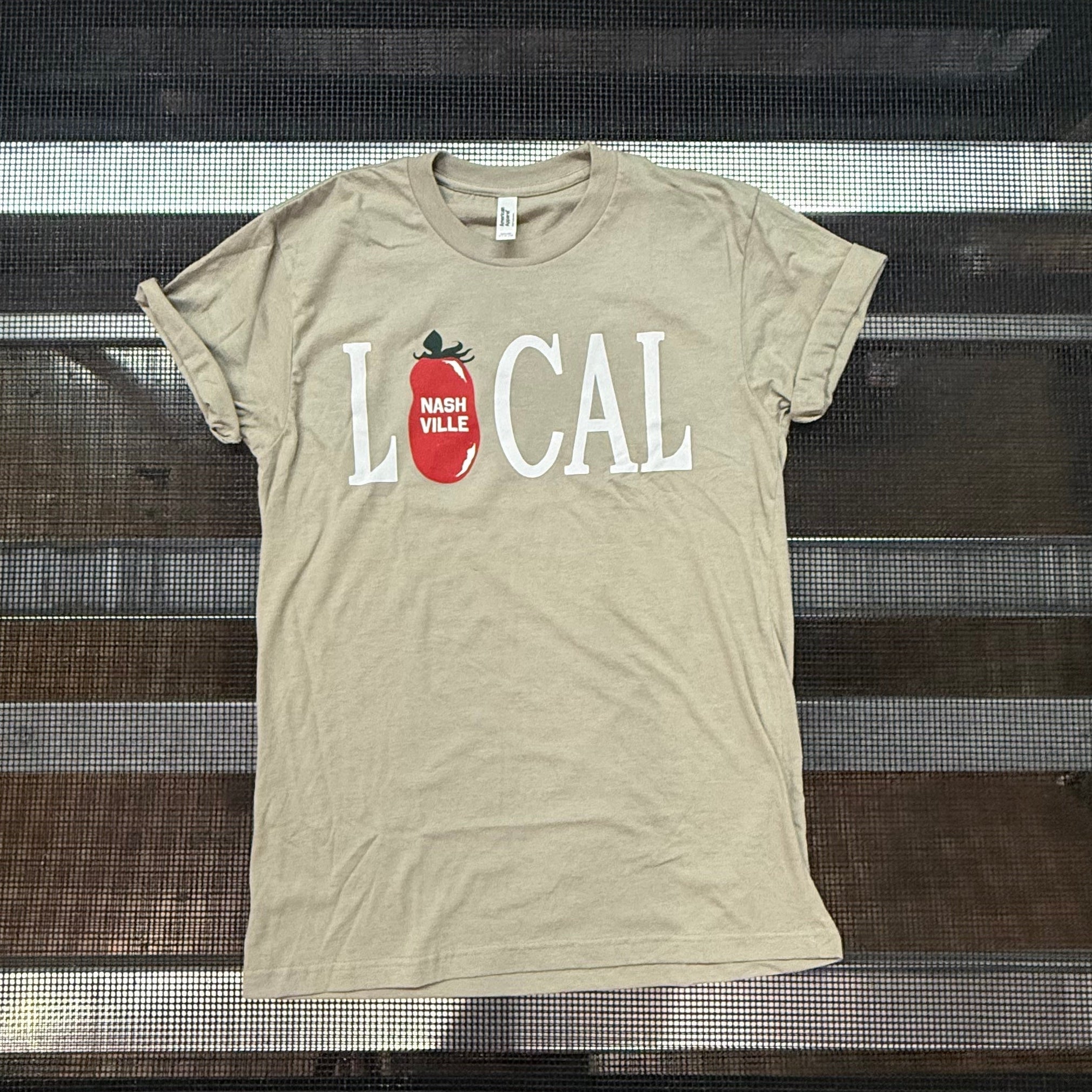 A beige graphic t-shirt with 'LOCAL' printed in white letters and a red tomato icon, laid flat on a black metal rack.