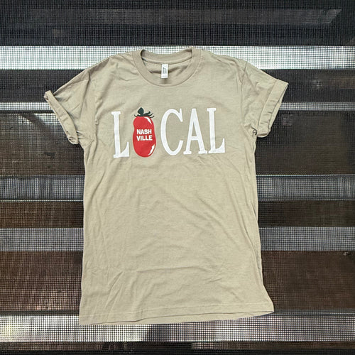 A beige graphic t-shirt with 'LOCAL' printed in white letters and a red tomato icon, laid flat on a black metal rack.
